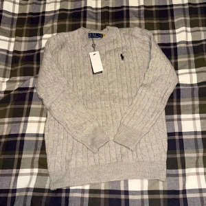 Grå kabelstickad tröja Polo Ralph Lauren - Kabelstickad grå tröja från Polo Ralph Lauren med klassisk rund halsringning och svart broderad logga på bröstet. Tröjan har ribbade muddar och är långärmad, perfekt för lager-på-lager under kyliga dagar.