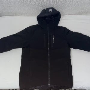 Svart dunjacka från Sail Racing - Svart dunjacka från Sail Racing med huva och hel dragkedja framtill. Jackan har två bröstfickor med dragkedja och diskret logga på bröstet och huvan. Perfekt för kalla dagar med sin vadderade design och stilrena look.