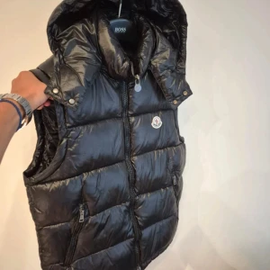 Svart dunväst från Moncler med huva - Snygg svart dunväst från Moncler med glansig finish och avtagbar huva. Västen har dragkedja framtill, två fickor med dragkedja och klassisk Moncler-logga på bröstet. Perfekt för lager-på-lager och streetstyle.