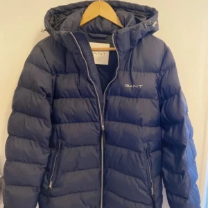 Mörkblå pufferjacka från GANT - Snygg mörkblå pufferjacka från GANT med huva och dragkedja framtill. Jackan har två fickor med dragkedja och broderad GANT-logga på bröstet. Perfekt för kyliga dagar och riktigt skön att bära.