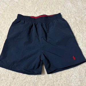 Marinblå badshorts Polo Ralph Lauren - Snygga marinblå badshorts från Polo Ralph Lauren med röd broderad logga på benet. Elastisk midja med röd insida och meshfoder. Klassisk passform och bakficka med lock. Perfekta för stranden eller poolen.