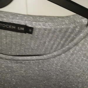 Glittrig ribbad tröja med knyt - Säljer en glittrig ribbad tröja från StockhLM i silver och svart. Tröjan har lång ärm, rund halsringning och en snygg knyt-detalj nertill. Perfekt för dig som vill sticka ut med lite extra glitter och stil. Storlek:M