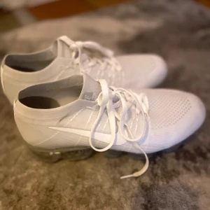 Nike Vapormax vita sneakers - Säljer ett par fräscha Nike Vapormax sneakers i helvitt med stickad ovandel och genomskinlig sula. Skorna har snörning och en sportig, modern look med tydlig Nike-logga på sidan. Perfekta för dig som gillar stilrena och bekväma sneakers.