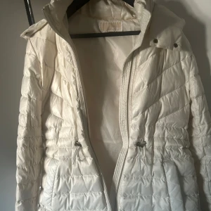 Vit dunjacka från Michael Kors - Snygg vit dunjacka från Michael Kors med quiltad design, huva och dragsko i midjan. Jackan har dragkedja framtill och diskret MK-logga på ärmen. Perfekt för kyliga dagar och ger en clean look med sin ljusa färg och stilrena detaljer. Använd en gång