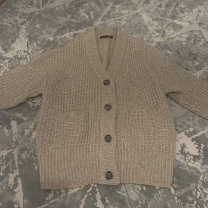 Beige stickad kofta med stora knappar - Mysig beige stickad kofta från Gina Tricot med grovstickat mönster, djup v-ringning och stora svarta knappar framtill. Koftan har två fickor och ribbade muddar vid ärmslut och nederkant. Perfekt för lager-på-lager och chill dagar.