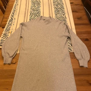 Grå långärmad klänning Vero Moda - En enkel och stilren grå klänning från Vero Moda med lång ärm och hög hals. Klänningen har en rak passform och är tillverkad i mjukt material som känns skönt mot huden. Perfekt för dig som gillar minimalistisk stil.