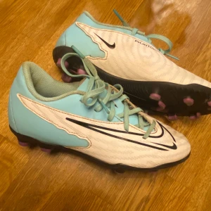 Nike Phantom GX fotbollsskor blå/vit - Nike Phantom GX fotbollsskor i ljusblått och vitt med svarta detaljer och rosa dobbar. Skorna har snörning och en snygg, sportig design med klassisk Nike-logga på sidan. Ovandelen är i syntetmaterial och sulan är anpassad för spel på gräs. De är knappt använda
