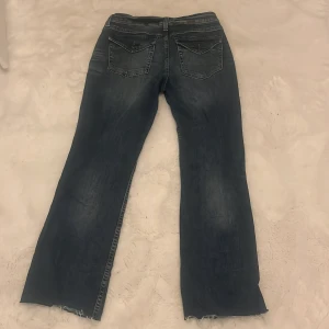 Blå bootcut jeans från H&M Divided - Snygga blå bootcut jeans från H&M Divided i storlek 42. Jeansen har slitna kanter nertill, klassiska bakfickor med lock och knapp, samt en tvättad look. Tillverkade i jeansmaterial med normal passform och låg midja.