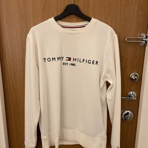 Vit sweatshirt från Tommy Hilfiger - Vit sweatshirt från Tommy Hilfiger med klassisk logga och text på bröstet. Tröjan har rund halsringning och långa ärmar. Perfekt för en clean och stilren look.
