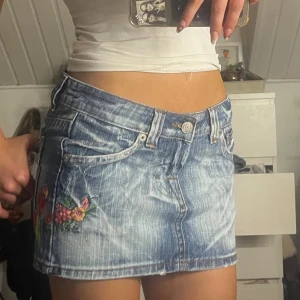 Broderad jeanskjol från Terranova - Säljer en snygg kort jeanskjol från Terranova med slitna detaljer och broderier av blommor, fjäril och papegoja. Kjolen har klassisk femficksdesign, låg midja och cool tvättad blå färg. Perfekt för dig som vill sticka ut med färgglada detaljer och street vibe.