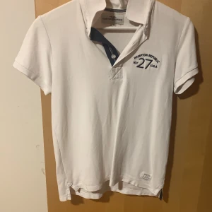 Vit pikétröja från Hampton Republic - Vit pikétröja från Hampton Republic med klassisk krage och korta ärmar. Broderad logga och text på bröstet samt snygga detaljer i mörkblått vid knäppningen. Perfekt för en clean och sportig stil.