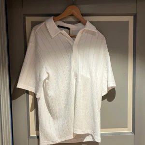 Två randiga skjortor med kort ärm - Säljer två skjortor med kort ärm och krage, en i vitt och en i mörkblått. Båda har diskreta vertikala ränder och knäppning framtill. Skjortorna har en avslappnad passform och är tillverkade i ett lätt, strukturerat material som känns svalt och skönt.