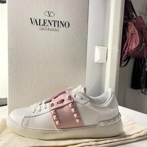 Valentino Rockstud sneakers vit/rosa - Snygga Valentino Garavani Rockstud sneakers i vitt skinn med ljusrosa detaljer och ikoniska nitar. Skorna har snörning, platt sula och rund tå. Perfekt för dig som vill ha en lyxig och trendig touch till din outfit.
