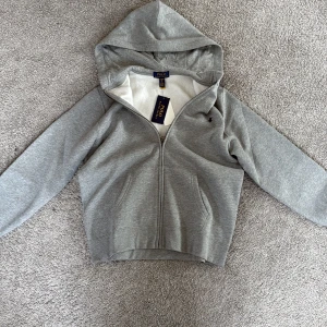 Grå hoodie från Polo Ralph Lauren XL - Snygg grå hoodie från Polo Ralph Lauren i storlek XL. Tröjan har dragkedja framtill, huva och två fickor. Klassisk broderad logga på bröstet och mjukt material som känns skönt mot huden. Perfekt för chill dagar eller när du vill ha en clean look.