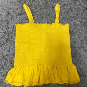 Gul volangtopp från KappAhl - Säljer en solgul linnetopp från KappAhl med smockad överdel och bred volang nertill. Toppen har smala axelband och en söt rynkad kant upptill. Perfekt för dig som vill ha en färgglad och somrig look.