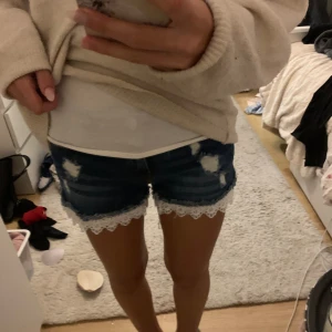Blå jeansshorts med spetskant - Snygga blå jeansshorts med slitna detaljer och vit spetskant längst ner. Klassisk femficksmodell med knapp och dragkedja framtill. Perfekt för dig som vill ha en cool och feminin look i sommar.