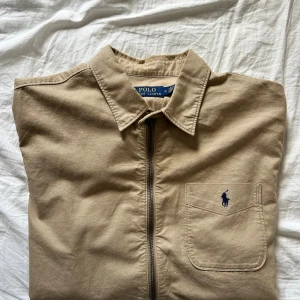 Beige overshirt från Polo Ralph Lauren - Säljer en beige overshirt från Polo Ralph Lauren. Plagget går givetvis att ha som en skjorta, men också som en jacka. Den har en dragkedja framtill och en bröstficka med loggan på. Ärmarna är långa och kan justeras med knappar vid ärmsluten. Denna overshirt, som egentligen inte har använts av mig så mycket, då den blivit för liten, tycker jag om väldigt mycket. Den fungerar med mer eller mindre allt, och passar i alla väder. Priset kan eventuellt diskuteras. 