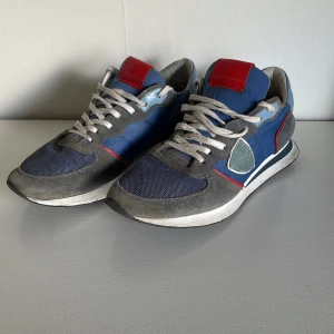 Philippe Model blå sneakers med mockadetaljer - Snygga sneakers från Philippe Model i blått med grå mockadetaljer och röda inslag. Skorna har vit sula, vita snören och en röd patch på plösen. Materialmix av mocka, textil och syntet ger en sportig och trendig look. Perfekta för dig som gillar streetstyle.