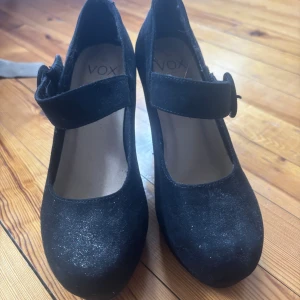 Svarta pumps med klack från Vox - Snygga svarta pumps med glitter från Vox med rund tå och bred klack. Skorna har en dekorativ rem över foten med en rund knäppning och är tillverkade i mocka för en mjuk och lyxig känsla. Perfekta för dig som vill ha en klassisk men ändå trendig look.     Priset +frakt