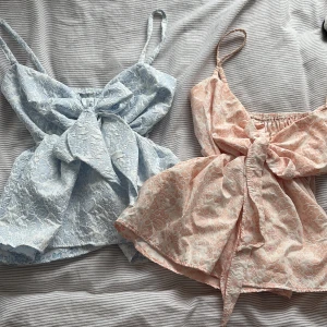 Blommiga linne-toppar med rosett - Två söta linne-toppar i pastellblått och ljusrosa med blommigt mönster och stor rosett framtill. Toppens material är lätt och har smala axelband samt smockad rygg för extra komfort.  Köpte topparna i Thailand och har använt de ca 1 gång. Säljer båda för 100kr (50kr styck )  ‼️(den rosa toppen har två fläckar, se sista bild )