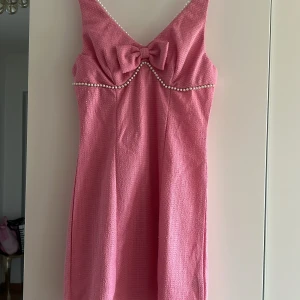 Rosa kort klänning med pärldetaljer - Supersöt rosa kort klänning med pärldetaljer längs urringningen och en stor rosett framtill. Klänningen har smala axelband, v-ringning och en figurnära passform. Materialet är strukturerat och ger en lyxig känsla. Perfekt för dig som vill sticka ut med en feminin look.