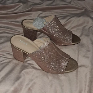 Glittriga klackskor från ICHI - Snygga glittriga klackskor från ICHI i en skimrande rose nyans. Skorna har öppen tå, bred klack och är täckta av glitter som ger en festlig look. Perfekta för dig som vill sticka ut med stil. Bekväm passform och platt sula framtill.