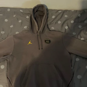 Grå Jordan PSG hoodie med gul logga - Snygg grå hoodie från Jordan x Paris Saint-Germain med stor gul PSG-logga på ryggen och mindre loggor på bröstet. Tröjan har huva med snörning och en stor magficka. Perfekt för dig som gillar streetwear och fotboll.