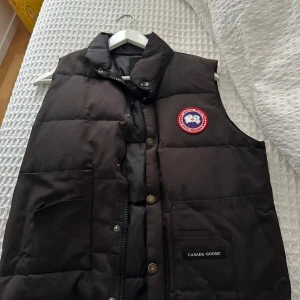 Svart dunväst från Canada Goose XS - Svart dunväst från Canada Goose med klassisk logga på bröstet och stora fickor framtill. Västen har hög krage, dragkedja och tryckknappar. Perfekt för lager-på-lager och riktigt snygg till streetwear-stilen.