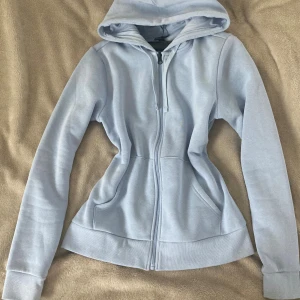 Ljusblå hoodie från Take Off - Ljusblå hoodie från Take Off i storlek S. Tröjan har dragkedja framtill, huva med snörning och två fickor. Den är figursydd med långa ärmar och ribbade muddar. Perfekt för chill dagar och enkel att matcha med jeans eller leggings.