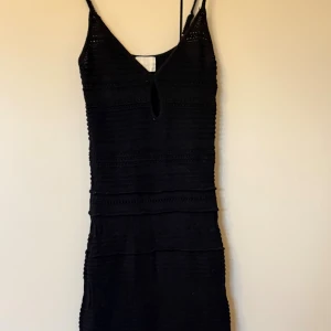 Svart stickad klänning från H&M - Supersnygg svart stickad klänning från H&M med smala axelband, v-ringning och liten öppning framtill. Klänningen har en figurnära passform och vågig nederkant, perfekt för dig som gillar en stilren men ändå unik look.