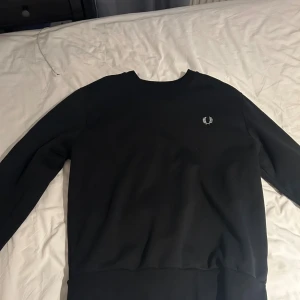 Svart Fred Perry sweatshirt - Svart sweatshirt från Fred Perry med klassisk broderad logga på bröstet och snygg detalj med loggor längs ena ärmen. Tröjan har rund halsringning och ribbade muddar. Perfekt för dig som gillar stilren streetwear.