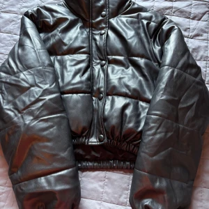 Svart pufferjacka, cropad i fejkskinn från Zara - Säljer en svart pufferjacka, cropad från Zara i fejkskinn. Jackan har hög krage, tryckknappar framtill och elastisk nederkant. Perfekt för dig som vill ha en cool och trendig look med lite edge. Jackan är vadderad och har en snygg, glansig finish.