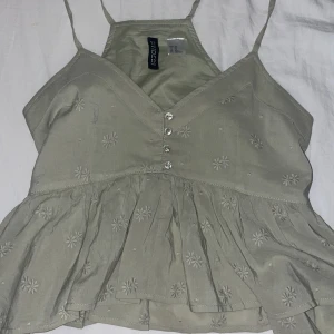 Grön broderad linnetopp med knappar - Supersöt Grön linnetopp från HM med broderade blommor och volang nedtill. Toppen har smala axelband, v-ringning och fyra pärlemorknappar framtill. Perfekt för varma dagar och ger en romantisk vibe till din outfit.