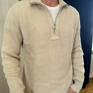 Beige stickad tröja med half zip - Säljer en beige stickad tröja med ribbad struktur och half zip-dragkedja vid halsen. Tröjan har lång ärm och en klassisk, avslappnad passform. Perfekt för lager-på-lager och enkel att matcha med jeans eller chinos.