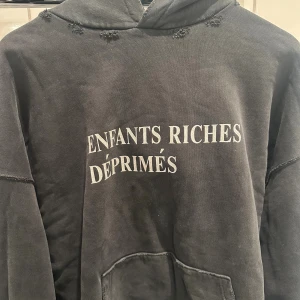 Svart hoodie Enfants Riches Déprimés - Cool svart hoodie från Enfants Riches Déprimés med vit text på bröstet. Oversized passform, huva och känguruficka framtill. Råa sömmar och slitna detaljer ger en edgy vibe. Perfekt för dig som gillar streetwear och statement-plagg.