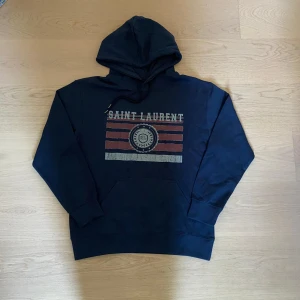 Mörkblå Saint Laurent hoodie  - Snygg mörkblå hoodie från Saint Laurent med tryck i rött och vitt framtill. Tröjan har huva med snörning och klassisk känguruficka. Tillverkad i mjuk bomull och perfekt för en chill och trendig look.