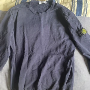Mörkblå sweatshirt från Stone Island - Säljer en mörkblå långärmad sweatshirt från Stone Island med den klassiska logotypen på vänster ärm. Tröjan har rund hals och är tillverkad i mjuk bomull, perfekt för dig som gillar streetwear och snygga detaljer. Säljer denna då den blivit för liten för mig och endast ligger i garderoben! Bara att hojta vid intresse!