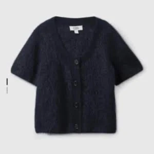 Mörkblå stickad kofta från COS - Kortärmad, mörkblå stickad kofta från COS med v-ringning och knappar framtill. Koftan har en mjuk och fluffig känsla, perfekt för lager-på-lager och en chill look. Enkel att styla till både jeans och kjol.