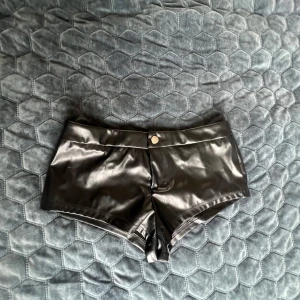 Svarta skinnshorts med knapp - Snygga svarta shorts i skinnimitation med låg midja och en silverfärgad knapp framtill. Modellen är tight och kort, vilket ger en cool och edgy vibe. Perfekta för dig som vill sticka ut och gillar trendiga detaljer.