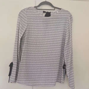 Vit och svart mönstrad blus med knyt - Säljer en snygg blus från Vero Moda i vitt och svart med grafiskt mönster. Blusen har långa ärmar med svarta knytband vid ärmsluten och en dekorativ knytning i nacken. Perfekt för dig som gillar stilrena detaljer och mönster.