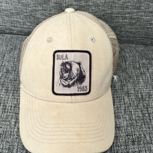 Beige keps från Bula med björnpatch Den kostar 500 igentligen  - Snygg beige keps från Bula med broderad patch framtill som visar en rytande björn och texten '1983'. Kepsen har klassisk böjd skärm och mesh-bakstycke för extra ventilation. Justerbar snapback för perfekt passform. Perfekt accessoar för en avslappnad stil.