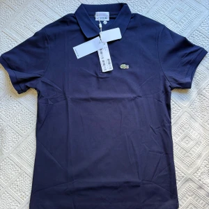 Mörkblå pikétröja från Lacoste - Klassisk mörkblå pikétröja från Lacoste med korta ärmar, krage och knappar framtill. Ikoniska broderade krokodilloggan på bröstet. Tillverkad i bomull för en skön och luftig känsla. Perfekt för en clean och sportig stil.