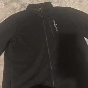 Svart vindjacka från Sail Racing - Svart vindjacka från Sail Racing med hög krage och dragkedja framtill. Jackan har en bröstficka med dragkedja och diskret logga på bröstet. Perfekt för dig som gillar stilren och sportig look.