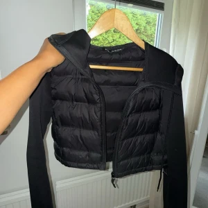 Svart croppad pufferjacka från Zara - Svart croppad pufferjacka från Zara med quiltad front och långa ärmar i slät polyester. Jackan har dragkedja framtill och en sportig vibe, perfekt för dig som gillar trendiga och stilrena plagg. Storlek S. 250 kr