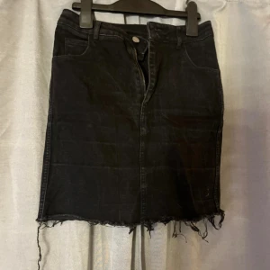 Svart jeanskjol från &Denim, strl 34 - Svart jeanskjol från &Denim i storlek 34. Kjolen är kort med rå, fransig fåll och klassiska jeansdetaljer som fickor fram och bak samt dragkedja och knapp. Perfekt för en edgy look med t-shirt eller hoodie.