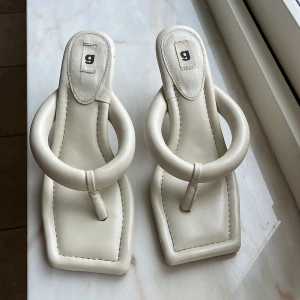 Beiga sandaler med klack från Ganni - Trendiga beiga sandaler från Gina med fyrkantig tå och chunky rundad rem. Skorna har en låg klack och är tillverkade i syntetmaterial med en cool, modern design. Perfekta för dig som vill sticka ut med en unik och stilren look.