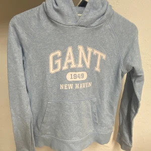 Ljusblå hoodie från GANT med tryck - Ljusblå hoodie från GANT med vit text 'GANT 1949 NEW HAVEN' på bröstet. Klassisk känguruficka framtill och huva. Mjuk och skön bomullsblandning, perfekt för chill dagar. Snygg och enkel stil som funkar året runt.