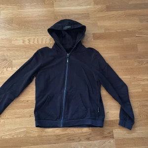 Svart hoodie från Plein Sport - Svart hoodie från Plein Sport med dragkedja framtill, huva och fickor. Snyggt röd Plein Sport-tryck på ryggen och diskret logga på dragkedjan. Tillverkad i mjuk bomull, perfekt för en sportig och avslappnad look.