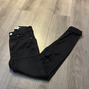 Svarta jeans med hål i knäna - Svarta jeans från lager 157 med hög midja och snygga slitningar på båda knäna. Klassisk femficksmodell med silverfärgade nitar och knapp. Jeansen har en rak passform och är tillverkade i ett stretchigt bomullsmaterial för extra komfort. Har alltid haft jeansen uppvikta så dom är inte slitna nertill, storlek XS.