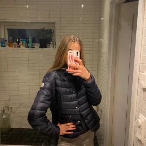 Navy Moncler puffjacka - Säljer en svart pufferjacka från Moncler med klassisk quiltad design och tryckknappar framtill. Jackan har Moncler-logga på ärmen och är lätt vadderad, perfekt för höst/ vår. Modellen är kortare och har två stora fickor framtill. Riktigt snygg och stilren jacka något stor för mig tror den passar en S eller M bäst🥰